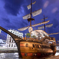 A wooden sailing ship with three masts docked at a riverside at night, under a cloudy sky, with lit buildings in the background.