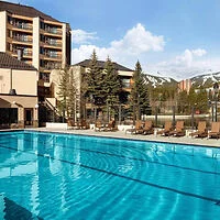 Outdoor pool with clear blue water, surrounded by lounge chairs. A multi-story building and pine trees are in the background against a partly cloudy sky.