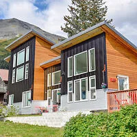 Modern two-story house with large windows, wooden and black paneling, set against a mountain backdrop and grassy lawn.