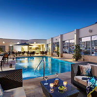 Rooftop pool area at dusk with chairs, tables, and lit candles, surrounded by modern buildings.