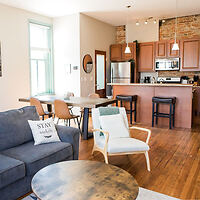 Open-concept living area with a gray couch, armchair, round coffee table, dining table, and a kitchen featuring wooden cabinets and a breakfast bar with stools.