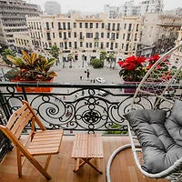 Balcony with a wooden chair, a small table, and a hanging cushioned chair overlooking a city square. Potted plants line the railing.