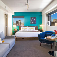 Hotel room with a king-size bed, blue accent wall, and modern furniture including a gray sofa and blue chair. Large windows offer a city view. A small glass table holds a tray with snacks.