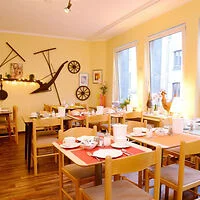 A cozy dining area with wooden tables and chairs, set for breakfast. Bright yellow walls are decorated with rustic farming tools and framed pictures near large windows.