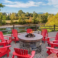 An outdoor seating area with red Adirondack chairs around a stone fire pit. Trees and a river are in the background.