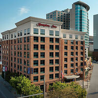 Exterior view of a Hampton Inn hotel, featuring a red brick facade and large windows, situated in an urban setting with surrounding buildings and a clear sky.