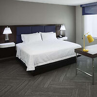 A hotel room with a large bed, two bedside lamps, a yellow desk lamp, and a window. The room has grey walls and flooring.