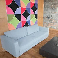 A light blue sofa sits in front of a colorful geometric wall. The room features wooden flooring and an exposed brick section.