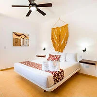 A neatly arranged bedroom with a double bed, decorative pillows, wall art, a ceiling fan, and soft lighting. A large, golden tapestry hangs above the bed.