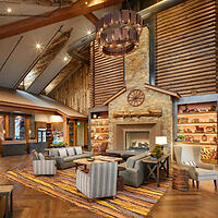 Spacious lodge interior featuring a stone fireplace, modern seating, wooden beams, and decorative shelving. Warm lighting enhances the cozy atmosphere.