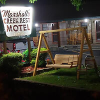 Motel exterior at night with a lit sign reading "Marshall's Creek Rest Motel." A wooden swing and chairs are situated on the grass nearby.