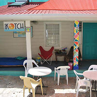 Outdoor patio with white chairs and tables. Red roof and teal door. A colorful post supports the canopy. "Kotch" sign hangs above.