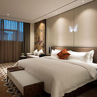 Modern hotel room with a large bed, decorative pillows, a bench at the foot, curtains, patterned carpet, and wall art. Soft lighting creates a cozy atmosphere.