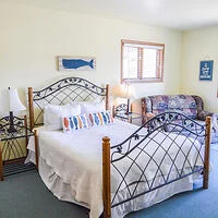 A bedroom with a wrought iron bed, white bedding, two nightstands with lamps, a couch, and minimal wall decor. A window lets in natural light.