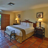 Hotel room with a king-sized bed, two nightstands with lamps, framed art on walls, and a bench at the foot of the bed. Tile flooring and wooden shutters complete the decor.