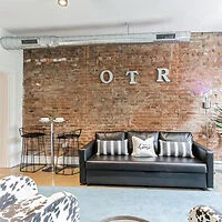 Loft-style room with exposed brick wall, metal ductwork, and "OTR" letters. Black leather sofa with striped pillows, cow-print chair, bar table, and stools.