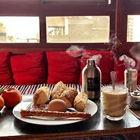 A breakfast scene with a plate of bread, fruit, and a boiled egg. A steaming glass of coffee and a thermos are on a wooden table with red cushions in the background.