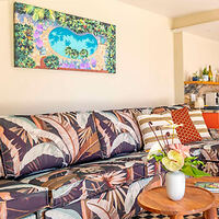 Colorful living room with a patterned sofa, tropical-themed cushions, and a wooden round table with a plant. A vibrant landscape painting hangs on the wall above the sofa.