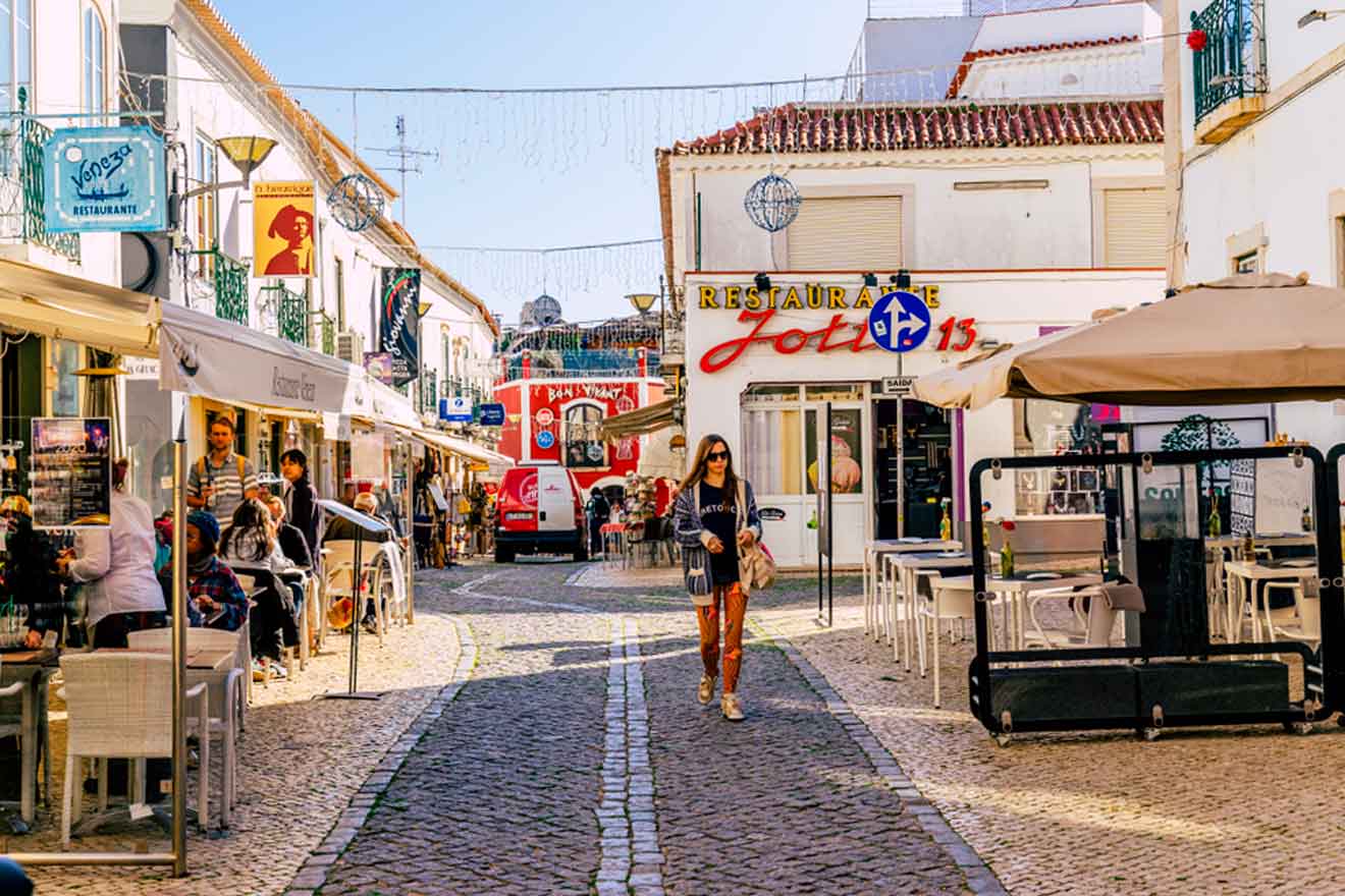 A pedestrian strolls along a cobblestone street lined with cafes and shops on a sunny day.