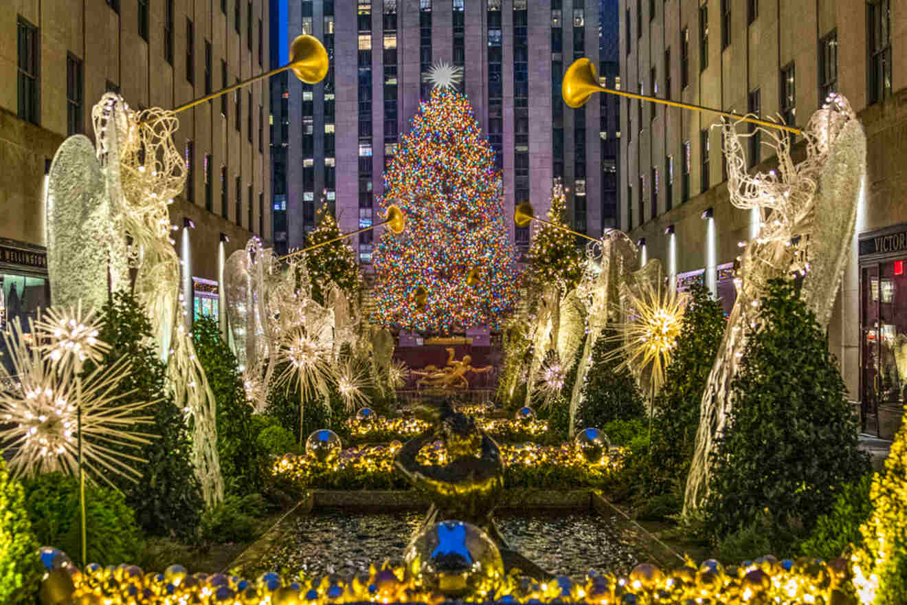 Illuminated Christmas tree and statues of angels with trumpets adorned with lights, surrounded by decorated bushes, in a city plaza at night.