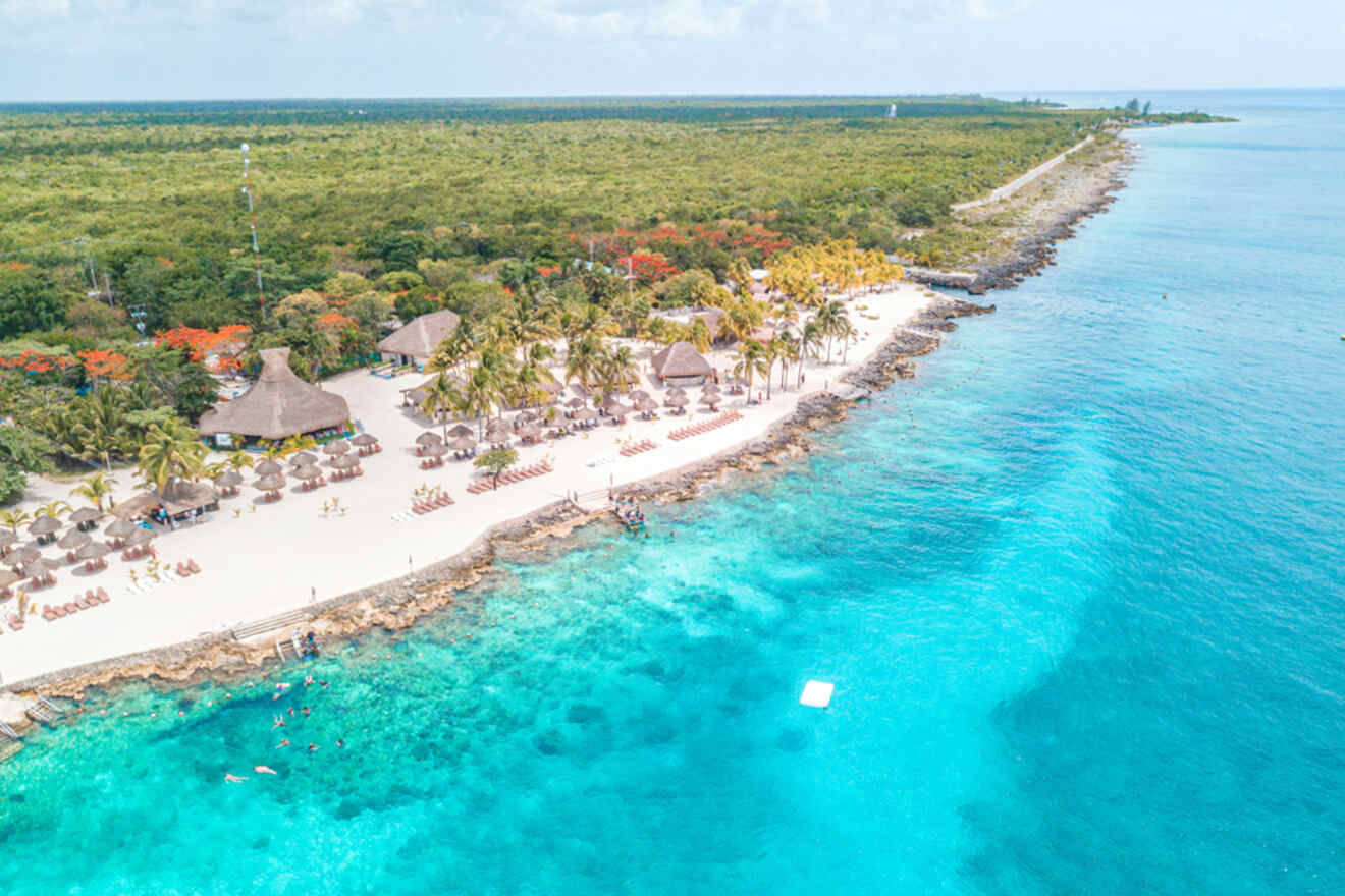 Aerial view of a tropical beach with thatched roof huts, white sand, and clear turquoise water. Lush greenery covers the land beyond the beach.