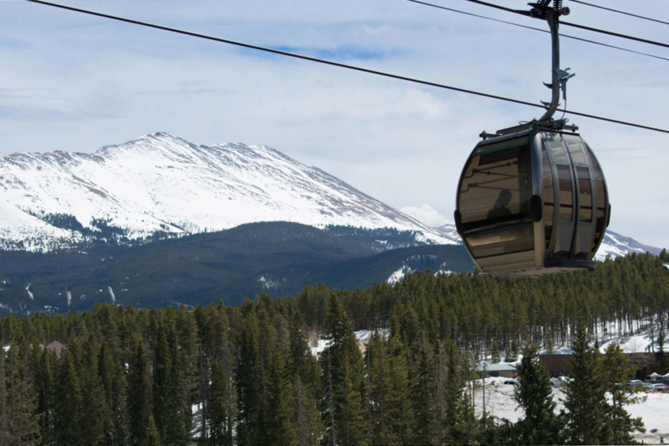 A gondola lift travels over a forested area with snow-covered mountains in the background.