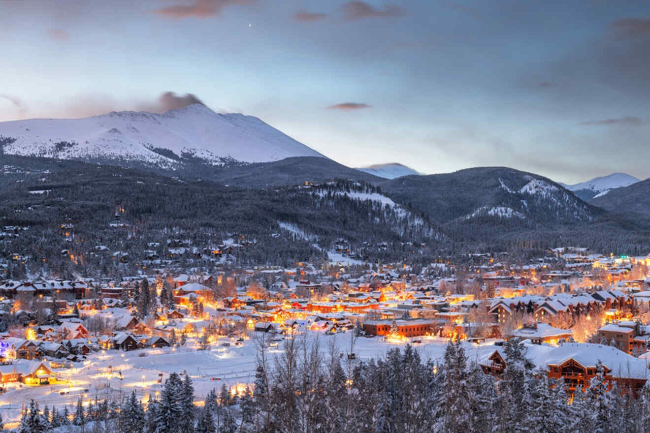 Snow-covered town with illuminated buildings at dusk, surrounded by mountains and trees.