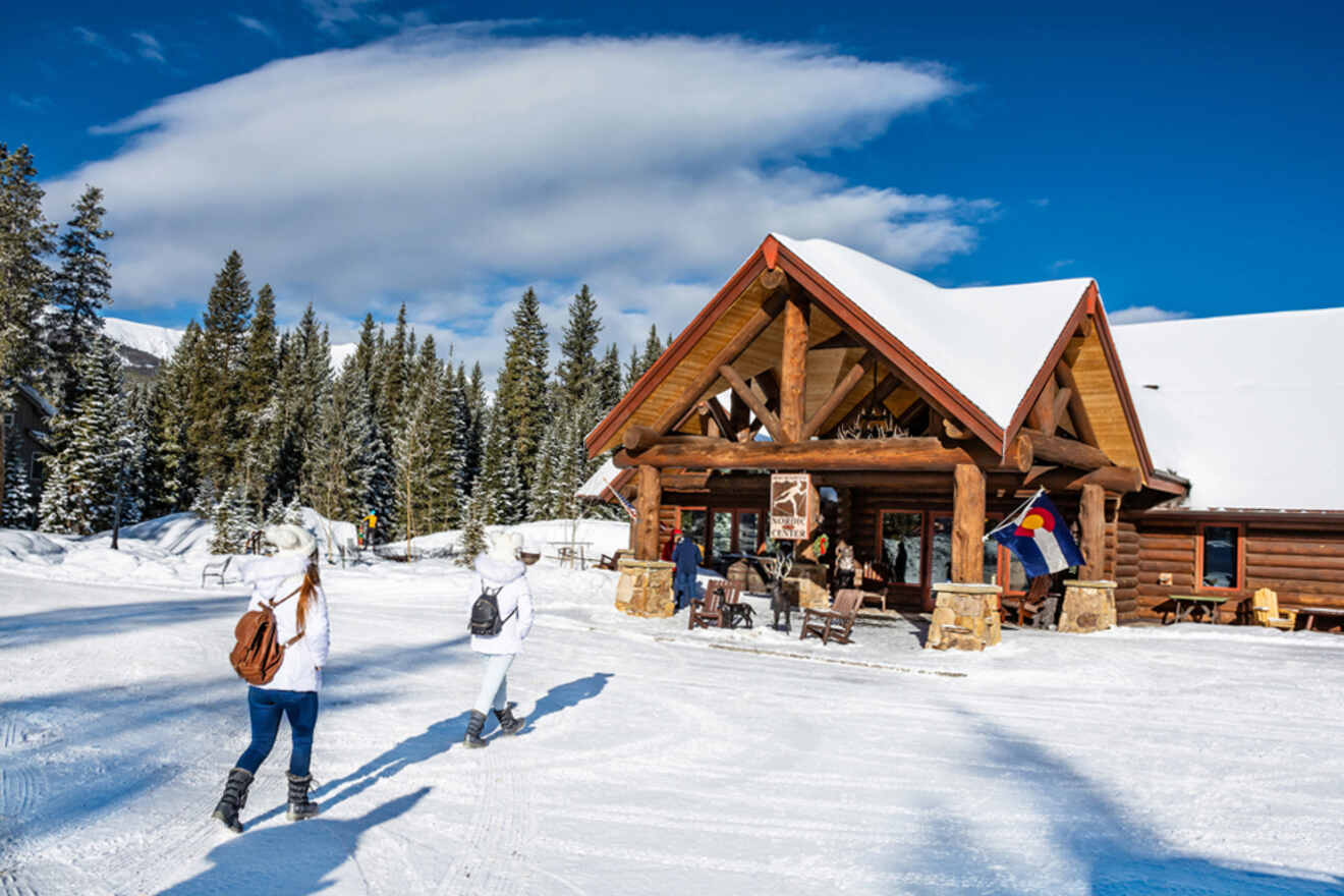 Two people walk towards a wooden lodge in a snowy landscape with pine trees and a clear blue sky.