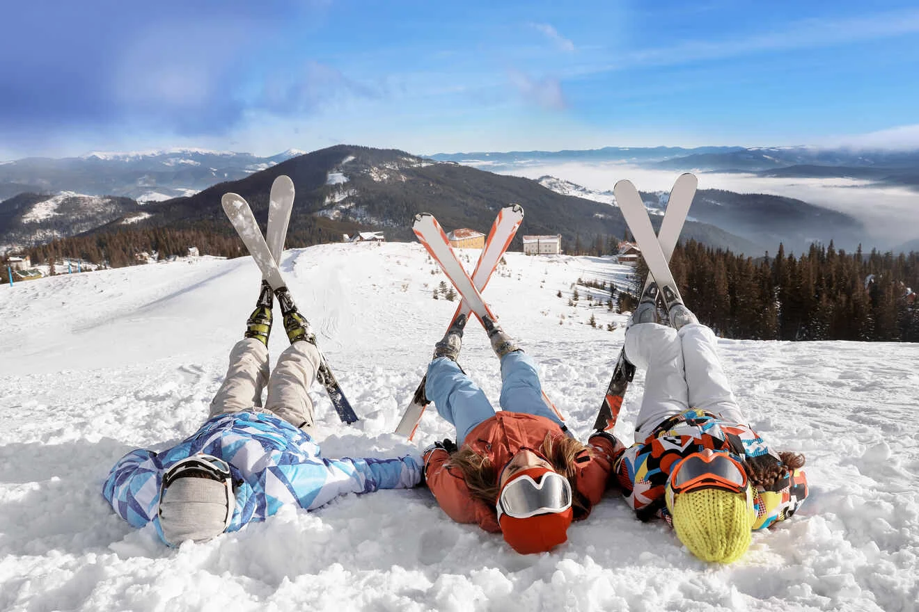 Three people in ski gear lie on their backs in the snow with crossed skis, overlooking a snowy mountain landscape.