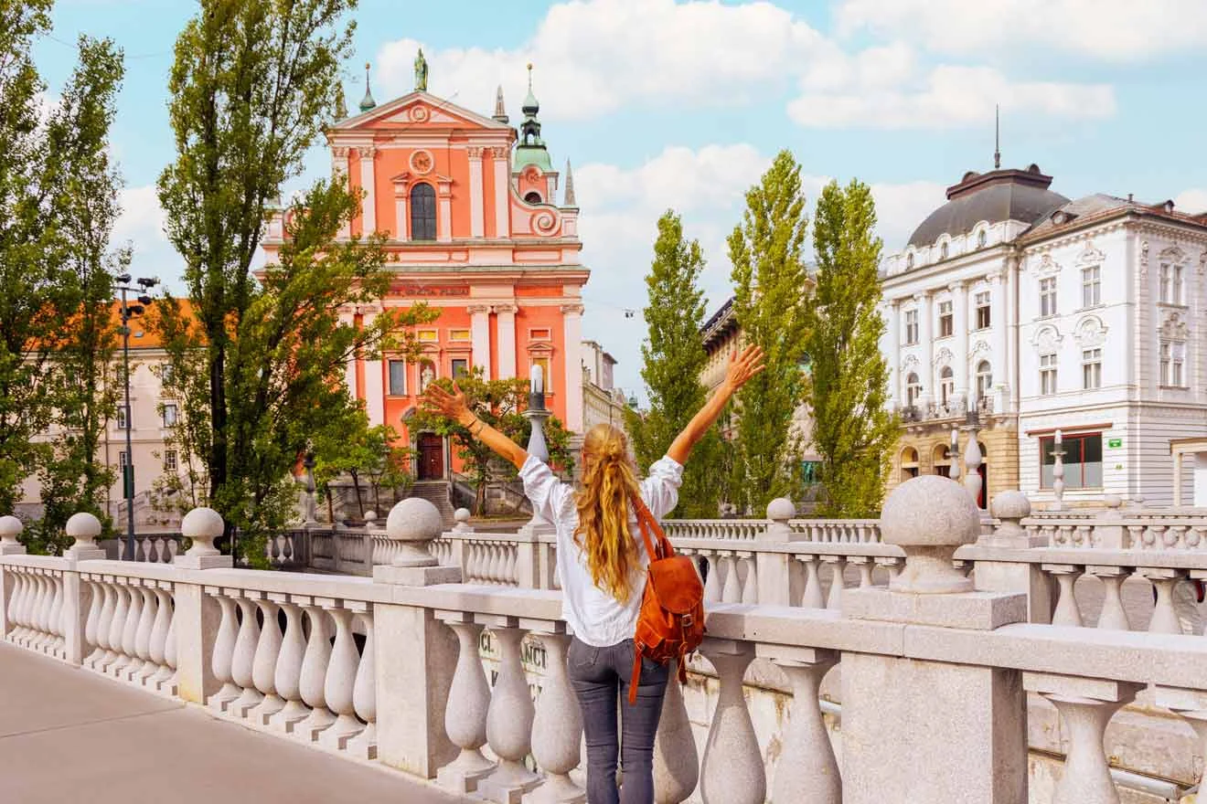 Person with arms raised stands on a bridge, facing a pink building with green spires and surrounding trees.