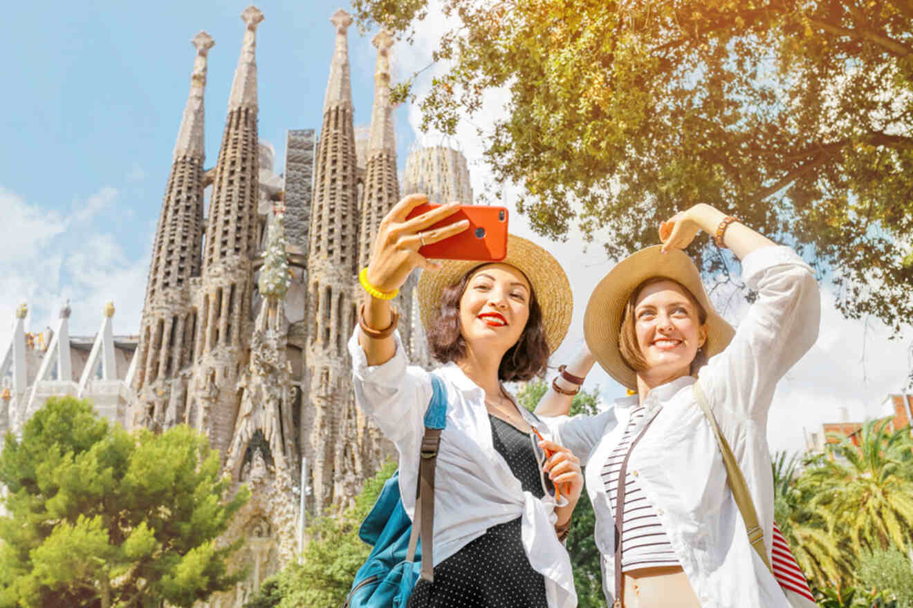 Two people take a selfie in front of the Sagrada Familia in Barcelona. They are wearing hats and smiling, with trees and the basilica's towers visible in the background.