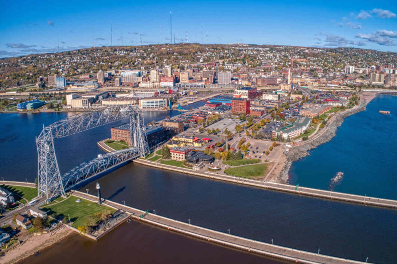 Aerial view of a city with a large lift bridge over a canal, surrounded by water and urban landscape. Hills and scattered clouds are visible in the background.