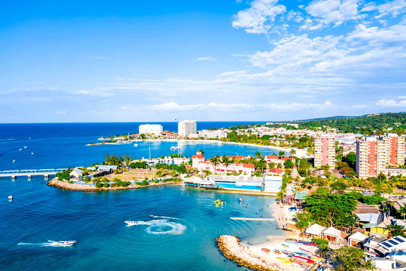 Aerial view of a coastal city with high-rise buildings, a marina, and beaches. The ocean is blue with several boats, and the sky is partly cloudy.