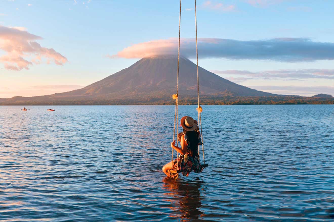 Person on a swing above water with a distant mountain and a partly cloudy sky in the background.