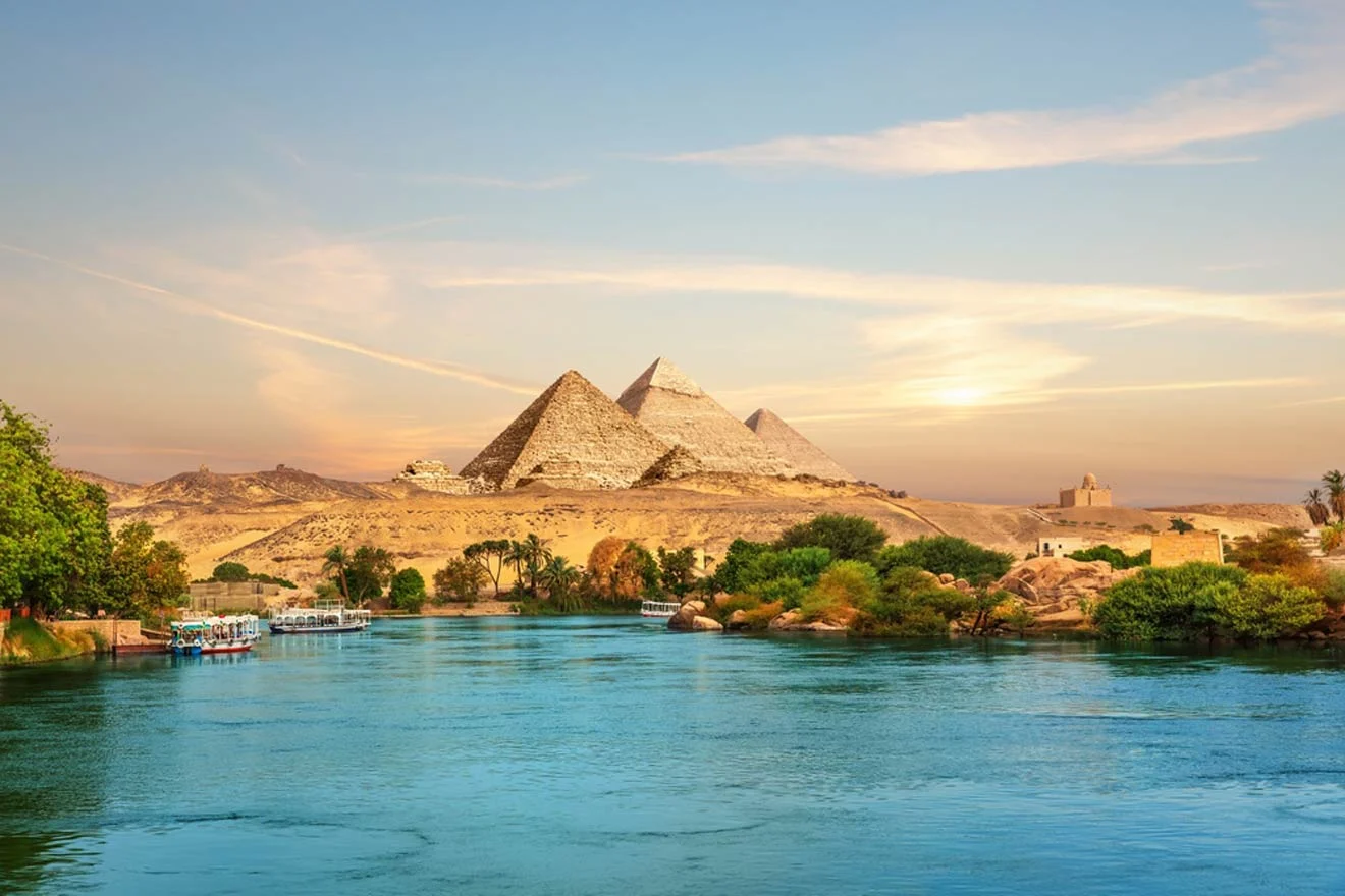 The pyramids of Giza, with the Nile River in the foreground, under a clear blue sky at sunset.