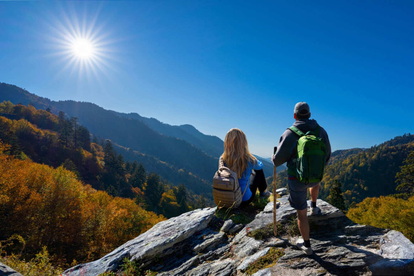 Two people with backpacks enjoy a scenic view from a rocky vantage point, overlooking forested mountains under a bright blue sky and sun.