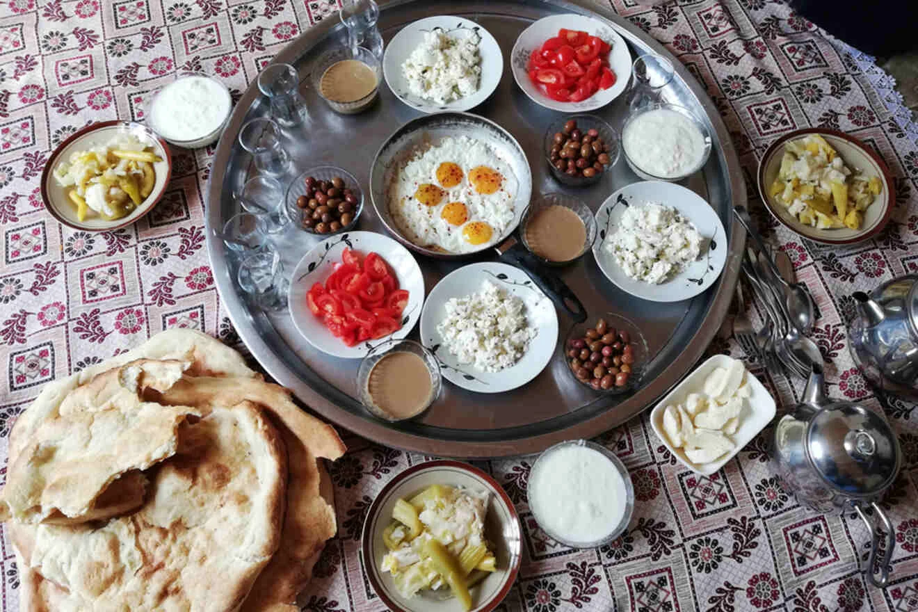 A table is set with flatbread, a large tray of dishes including rice with eggs, tomatoes, yogurt, pickles, olives, and drinks, all placed on a patterned tablecloth.