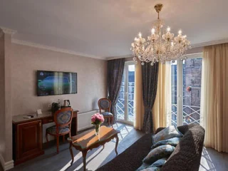 A living room with a chandelier, sofa, wooden coffee table with flowers, a wall-mounted TV, and large windows with curtains.