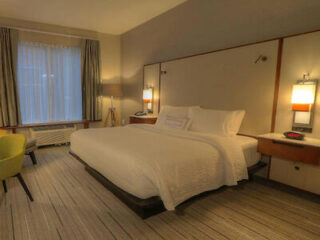 A hotel room with a king-size bed, white linens, two side tables with lamps, a window with curtains, a green chair, and a large wall mirror.