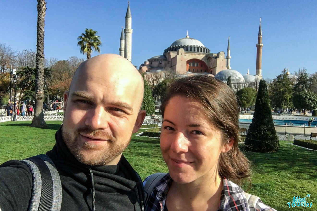 The writer of the post with her husband take a selfie in a park with a historic domed building and minarets in the background.