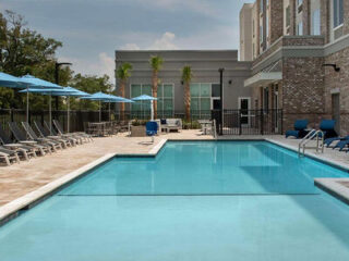 Outdoor swimming pool area with lounge chairs, blue umbrellas, and brick building background on a sunny day.