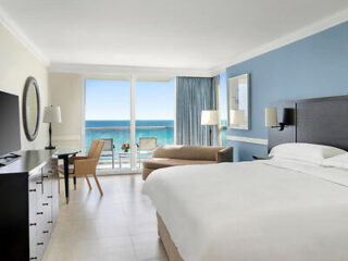 Hotel room with a large bed, a desk, and a chair. There's an ocean view through a sliding glass door leading to a small balcony with patio furniture.