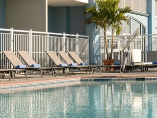 Lounge chairs lined up beside an outdoor pool with a metal fence and a potted palm tree in the background.