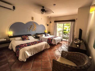 A hotel room with two beds, decorative blue headboard, and wall art. There's a flat-screen TV, wicker chair, and glass door leading to a balcony with greenery visible outside.