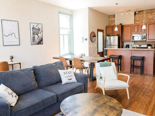 Open-concept living room and kitchen with a blue sofa, armchair, dining table, and bar stools. Decor includes wall art and cushions. Natural light from a large window brightens the space.