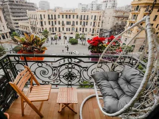 Balcony view with a wooden chair, small table, and hanging chair overlooking a city square with buildings and trees.