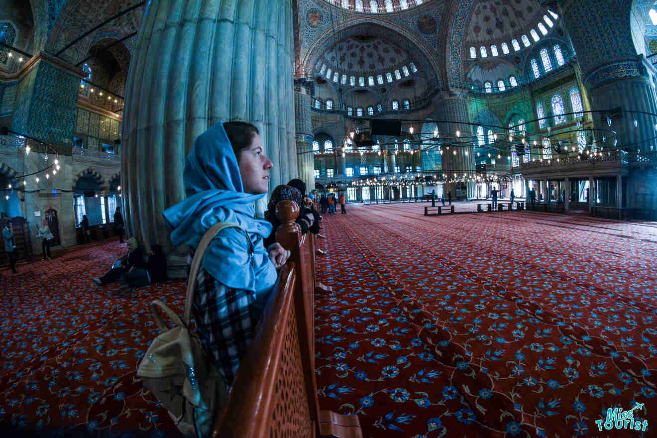 The writer of the post in a blue headscarf stands inside a large, ornately decorated mosque with red patterned carpet and domed ceilings.