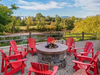 An outdoor seating area with red Adirondack chairs around a stone fire pit. Trees and a river are in the background.