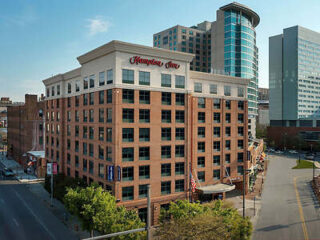 A multi-story brick Hampton Inn hotel, with a modern glass building in the background and a city street in the foreground.