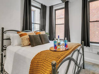 A cozy bedroom with a metal bed frame, white bedding, and black curtains. A mustard throw and pillows are on the bed, with a tray holding drinks and glasses. Brick walls are visible through windows.